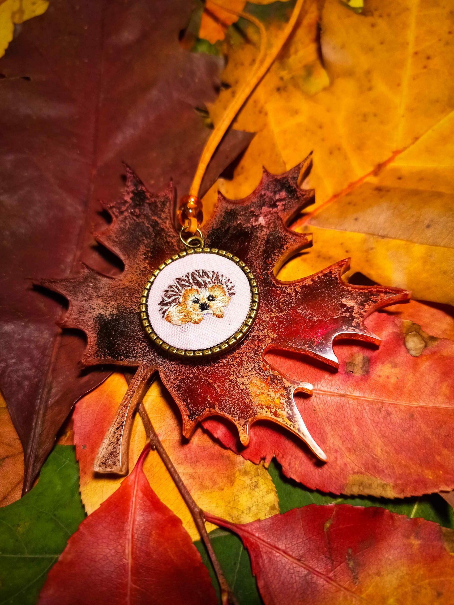 Medalion Jeżyk. Naszyjnik na skórzanym rzemieniu ozdobionym dwoma koralikami w kolorze żółtym i pomarańczowym. Haft z wizerunkiem jeża umiejscowiony jest w bazie ze stali nierdzewnej w kolorze brązu.