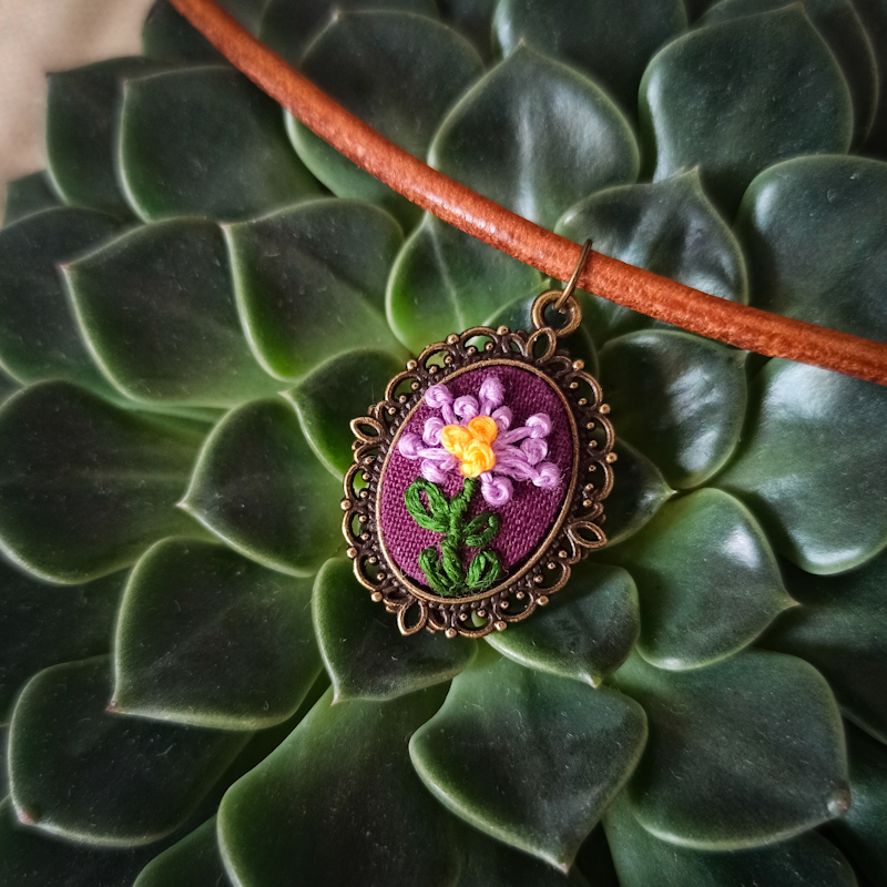 Naszyjnik Aster z haftem miniaturowym - fioletowym kwiatem Astra Alpejskiego.Haft jest przestrzenny. Kwiat wystaje około 1,5 mm ponad powierzchnię tkaniny, na której jest wyszyty. Owalna baza wisiorka w kolorze brązu z ozdobnymi detalami. Rzemień skórzany.